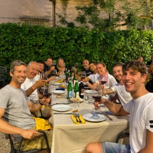 Un gruppo di persone sorridenti sedute a un tavolo all'aperto con bicchieri di vino e piatti.