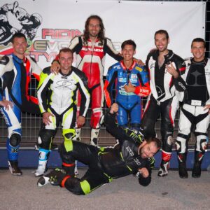 Gruppo di sei motociclisti in tute da corsa, posando insieme in un ambiente di gara.