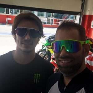 Due uomini sorridenti indossano occhiali da sole colorati, uno con logo Monster, in un ambiente di pista motociclistica.