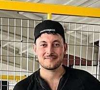 Uomo con cappellino nero e maglietta scura, sorridente, in un ambiente interno con recinzione metallica.