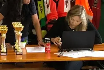 Holly firma documenti davanti a un laptop nero su un tavolo con coppe, mentre sullo sfondo si vedono tute arancioni e verde